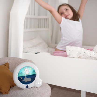 Pabobo Kid'Sleep Essential przy łóżku, w tle dziewczynka przeciągająca się po przebudzeniu.